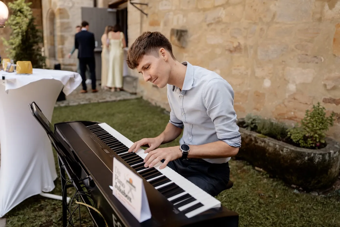 Silas spielt im Innenhof einer Hochzeitsfeier am Klavier, vorne das Schild der Piano Jukebox